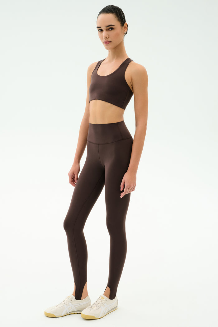 A woman stands in a studio wearing the SPLITS59 Sara Airweight Bra in Dark Chocolate with matching high-waisted stirrup leggings—ideal for Pilates or hot yoga—and finishes the look with white sneakers.