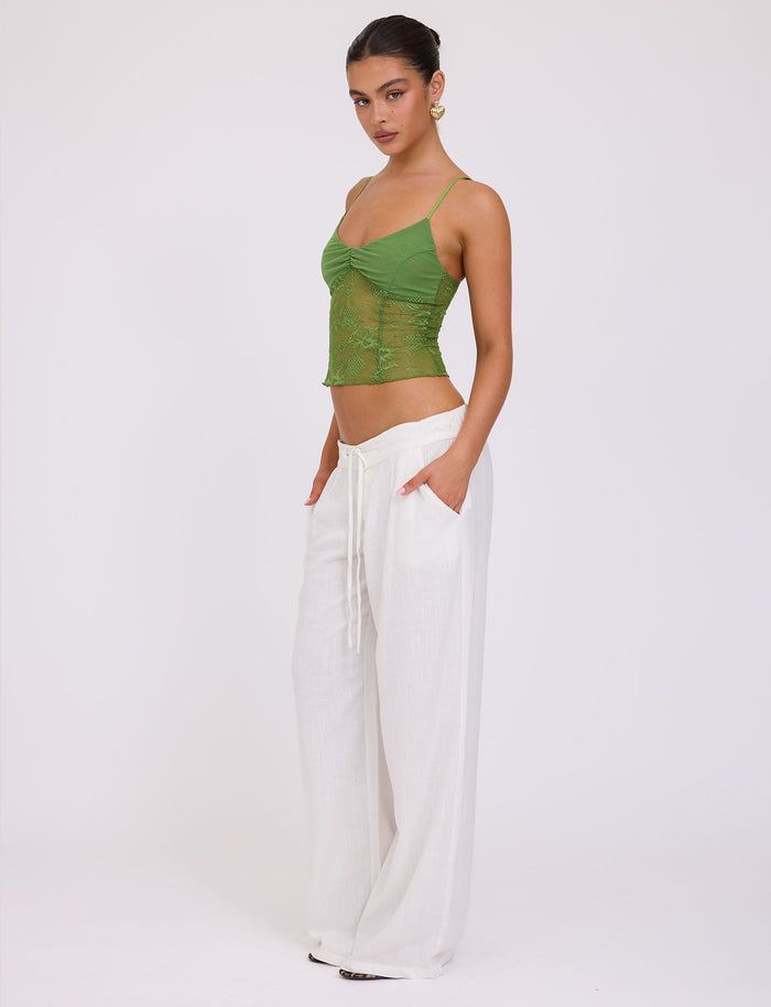 A woman stands against a white background, wearing the SASKIA TOP - GREEN from TIGERMIST paired with white wide-leg pants, her hands in her pockets. She has her hair tied back and is looking towards the camera with a neutral expression.