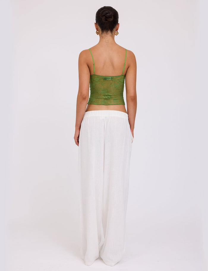 A person with their hair in a bun is standing and facing away from the camera, showcasing an ensemble that could be straight out of a TIGERMIST collection: the SASKIA TOP in green paired with loose, white pants. The background is white and plain.