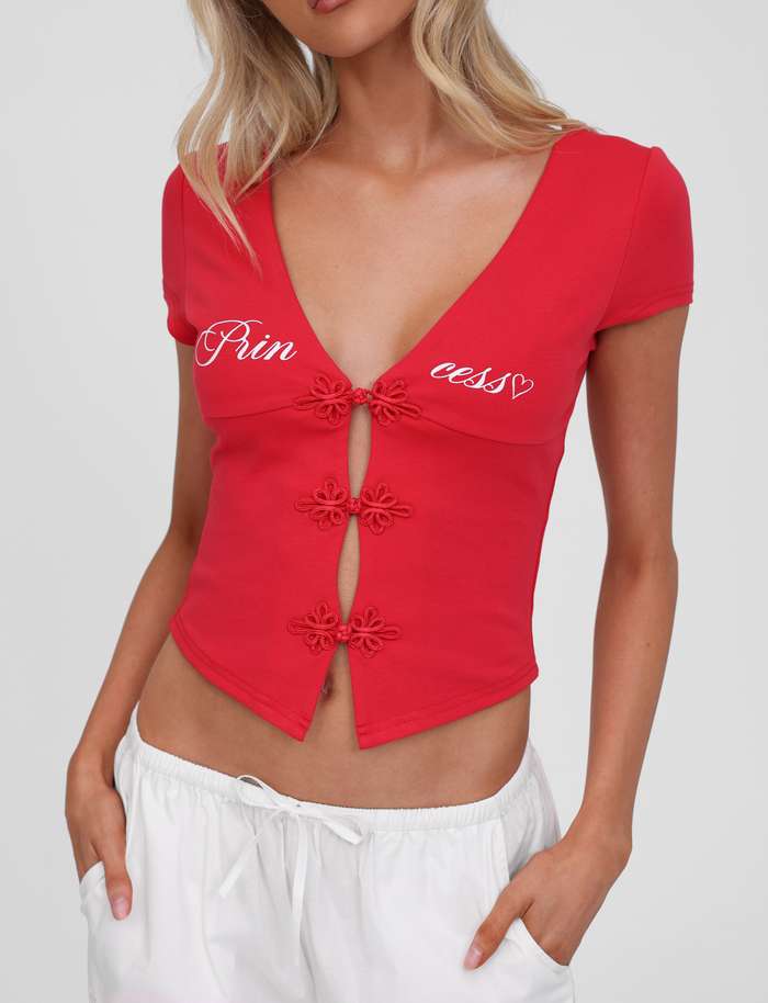A woman wears the TIGERMIST SAVINA TOP in red, featuring embroidered button details and a white "Princess" graphic print in cursive, paired with white pants. Her face is partially out of frame.