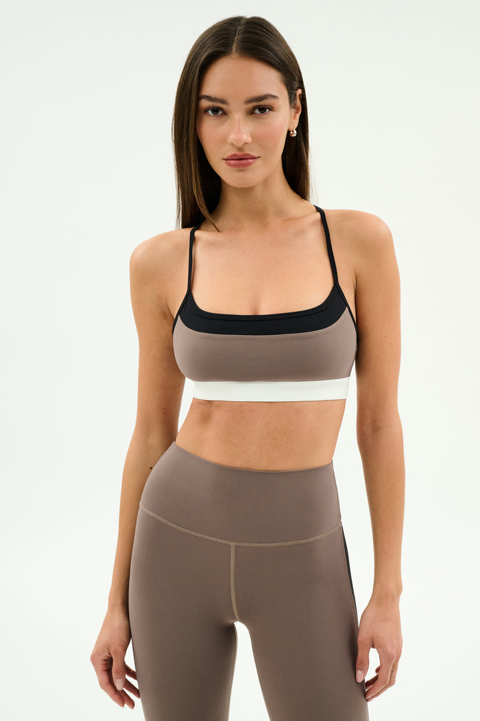 A woman models the SPLITS59 Scottie Airweight Bra in Lentil/Black, featuring black and white accents and matching high-waisted leggings, made from a soft nylon-spandex blend, against a plain white background.