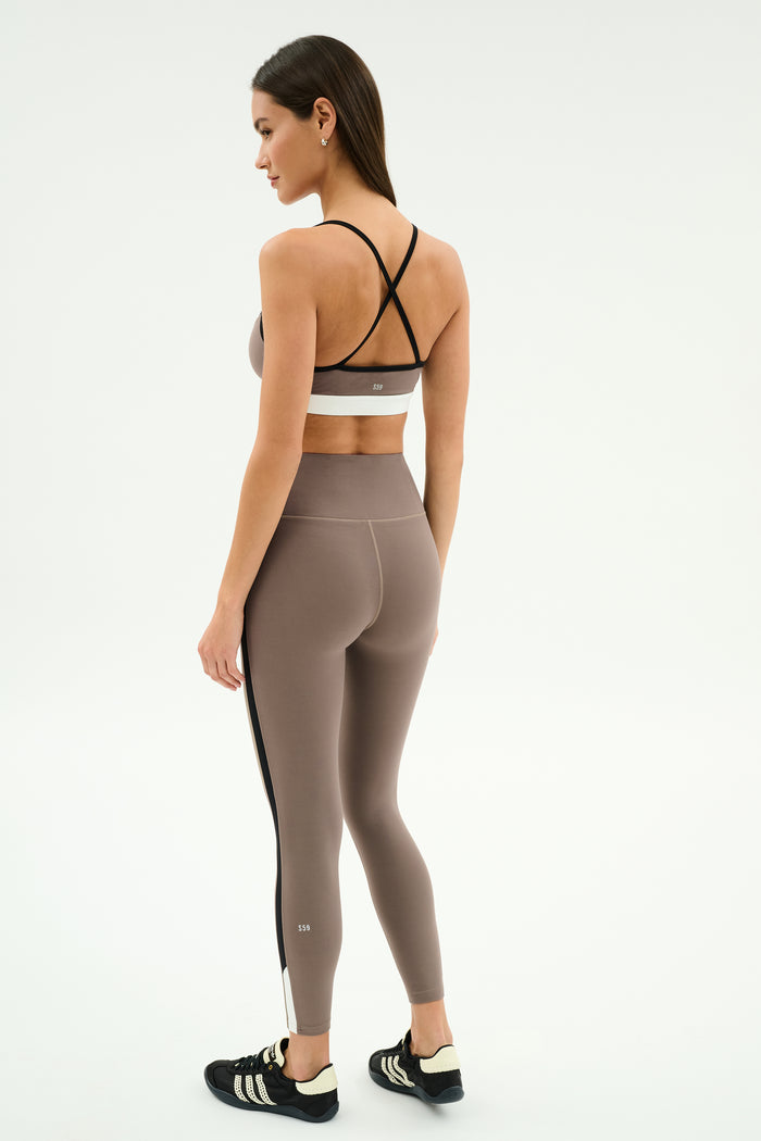 A woman faces away in the SPLITS59 Scottie Airweight Bra in Lentil/Black with matching high-waisted leggings and black athletic shoes, set against a plain background.
