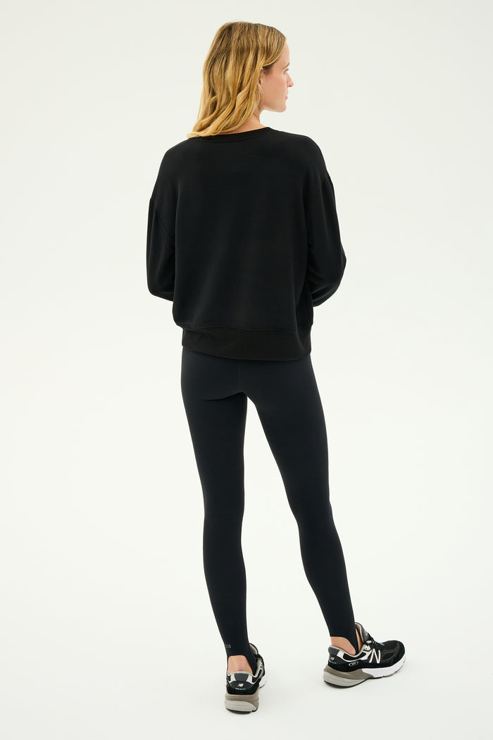 A person is standing against a neutral background, wearing the Sonja Fleece Sweatshirt in black crafted from ultra-luxe fleece by SPLITS59, paired with leggings and sneakers. The outfit effortlessly embodies comfort and style, exemplifying the MADE IN LOS ANGELES quality.