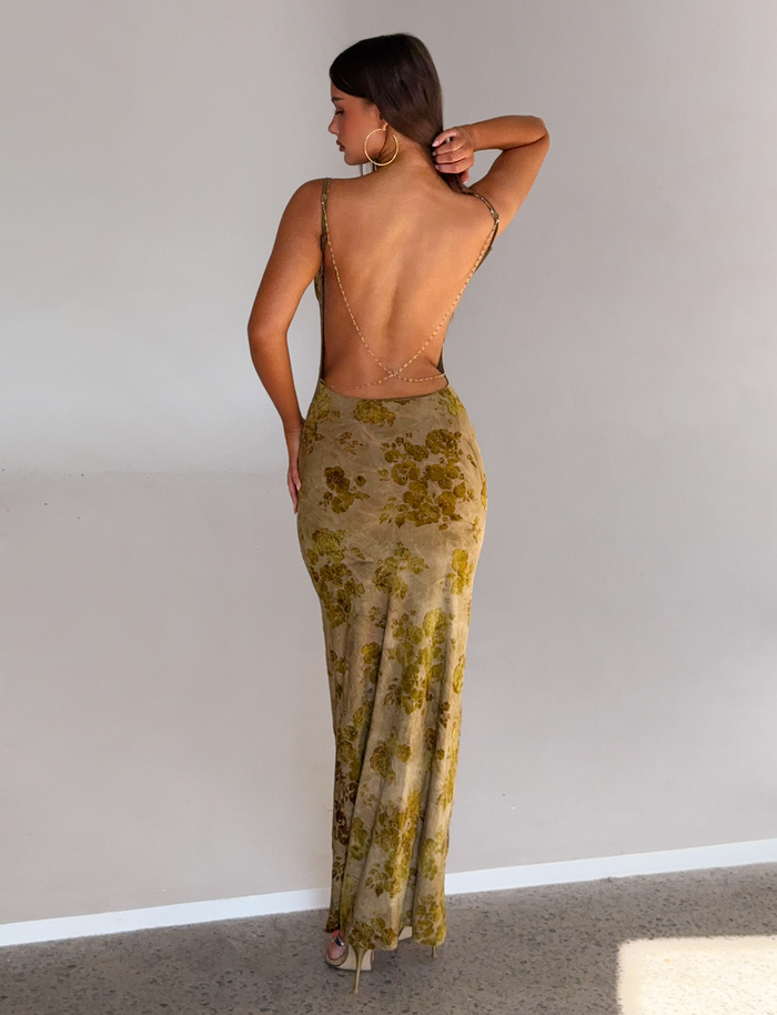 A woman stands with her back to the camera, wearing the TIGERMIST SPARROW MAXI DRESS in Green Burnout—a form-fitting, floor-length dress with gold chain straps and a yellow floral pattern—touching her hair against a plain light wall.