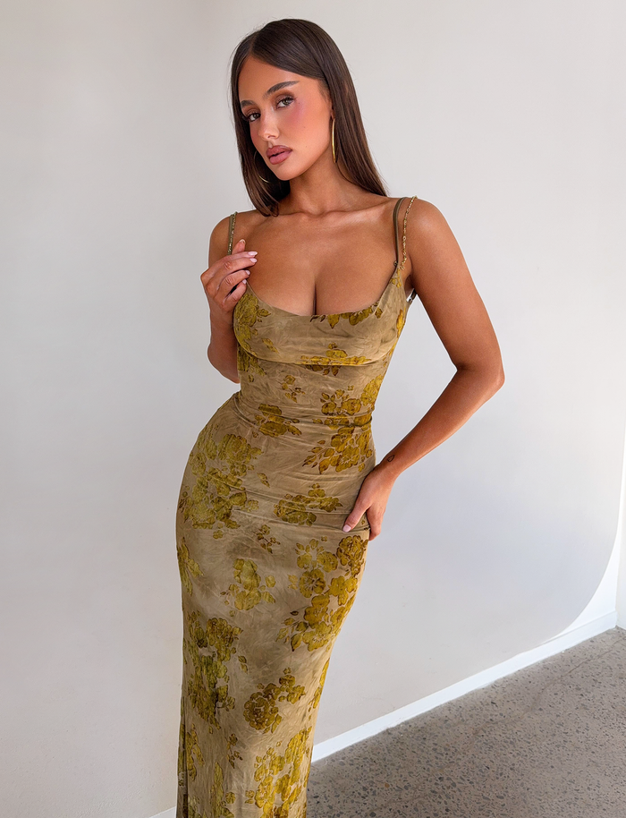 A woman with long brown hair poses indoors in the TIGERMIST SPARROW MAXI DRESS - GREEN BURNOUT, a fitted green floral dress with thin straps, standing against a white wall and looking slightly to the side.