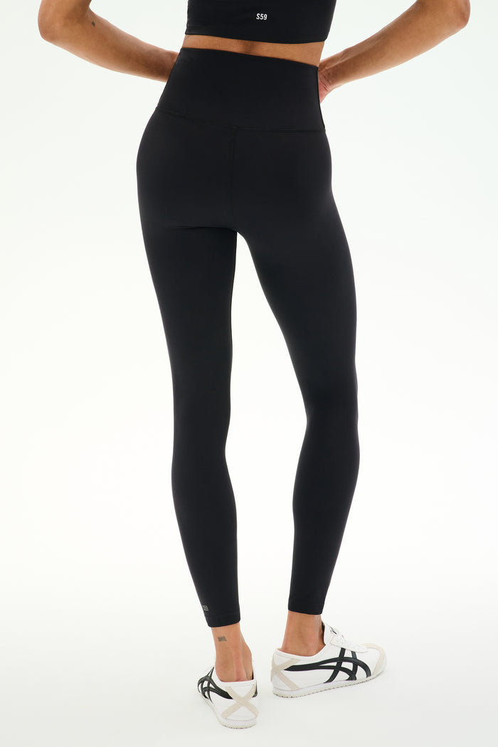 A person is shown from the back standing against a plain white background, wearing SPLITS59 Sprint High Waist Rigor 7/8 leggings in black made from durable Rigor fabric, paired with white athletic shoes.