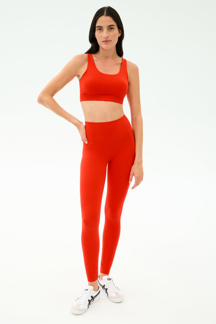 A woman stands against a plain background wearing the SPLITS59 Sprint Rigor Bra in Lotus Red with matching high-waisted leggings, ideal for multi-sport performance, and paired with white sneakers.