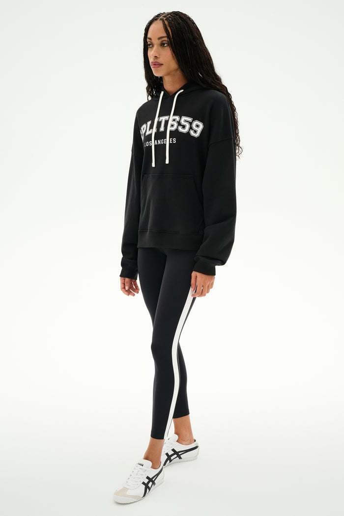 A woman wearing a black SPLITS59 Stella French Terry Oversized Hoodie with "Splits59 Los Angeles," black leggings with white stripes, and white sneakers stands against a white background.