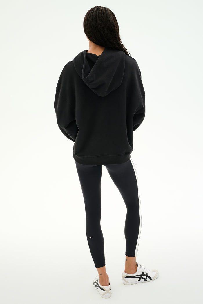 A person stands with their back to the camera, wearing the SPLITS59 Stella French Terry Oversized Hoodie in black, paired with black leggings featuring a white stripe and white sneakers, set against a plain white background.
