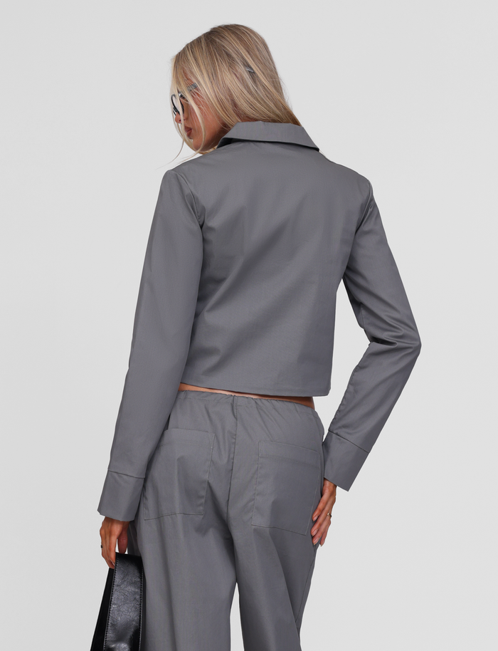 A woman with long blonde hair wears the TIGERMIST TYSHA JACKET in charcoal, paired with matching trousers, standing with her back to the camera while holding a black handbag and wearing sunglasses.