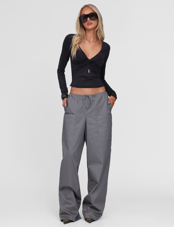 A woman in dark sunglasses, a black long-sleeve deep V-neck top, and TIGERMIST TYSHA PANT - CHARCOAL stands confidently with her hands in her pockets against a plain white background.