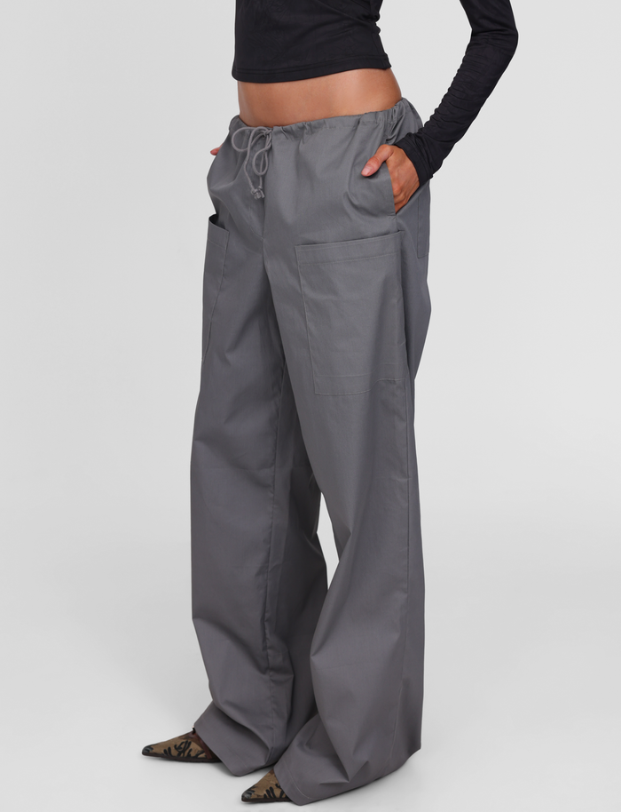 A person wears TIGERMIST’s TYSHA PANT in charcoal—wide leg cargo pants with a drawstring waist and large front pockets—styled with a cropped black long-sleeve top and pointed shoes, set against a plain white background.