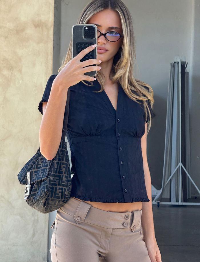 A woman with long blonde hair wears sunglasses, beige pants, and the TIGERMIST VALERIE SHORT SLEEVE SHIRT in black. She takes a mirror selfie with her phone and carries a black designer handbag featuring an "F" pattern.