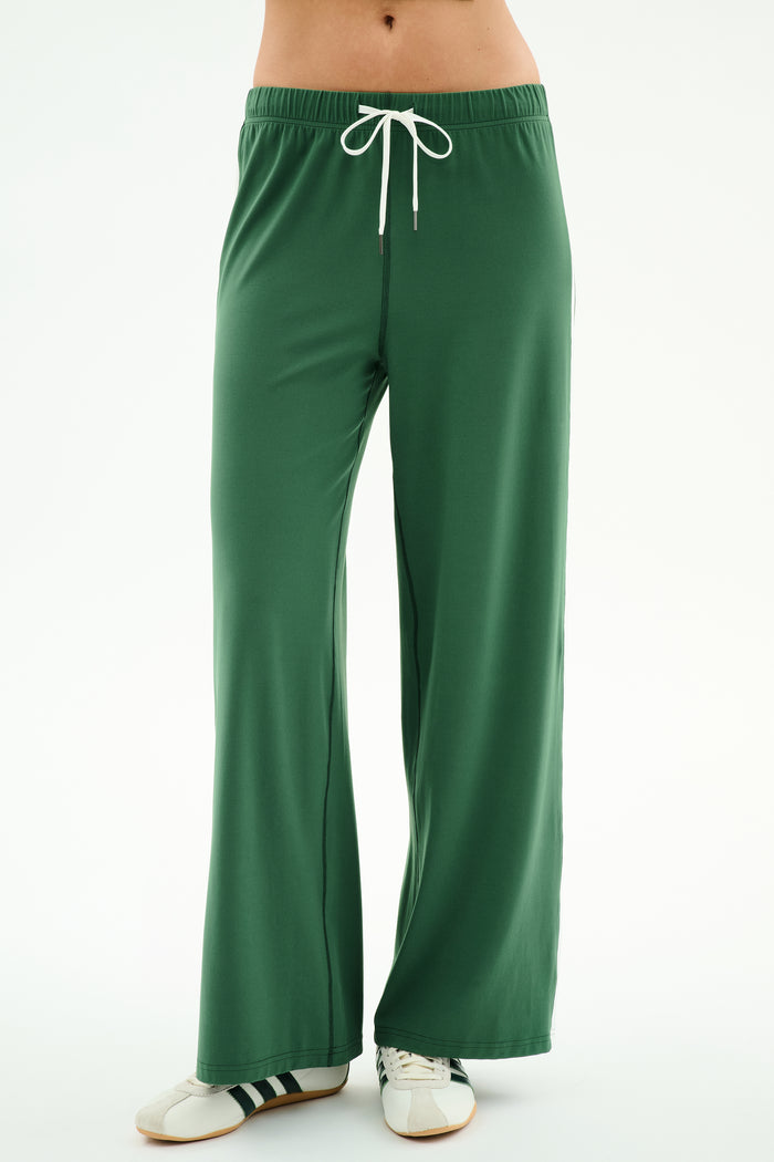 A person wearing SPLITS59 Willow Airweight Wide Leg pants in Arugula/White and white sneakers with black stripes stands against a plain white background.