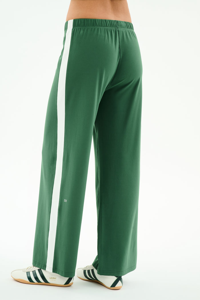 A person models SPLITS59’s Willow Airweight Wide Leg pants in Arugula/White with white sneakers featuring black stripes, standing against a white background.