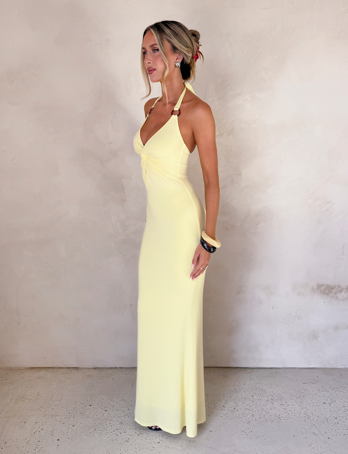 A woman stands sideways against a light textured wall wearing the TIGERMIST ZENEVA MAXI DRESS in Lemon. Her hair is styled up with loose strands framing her face, and she accessorizes with bracelets, looking calmly ahead.