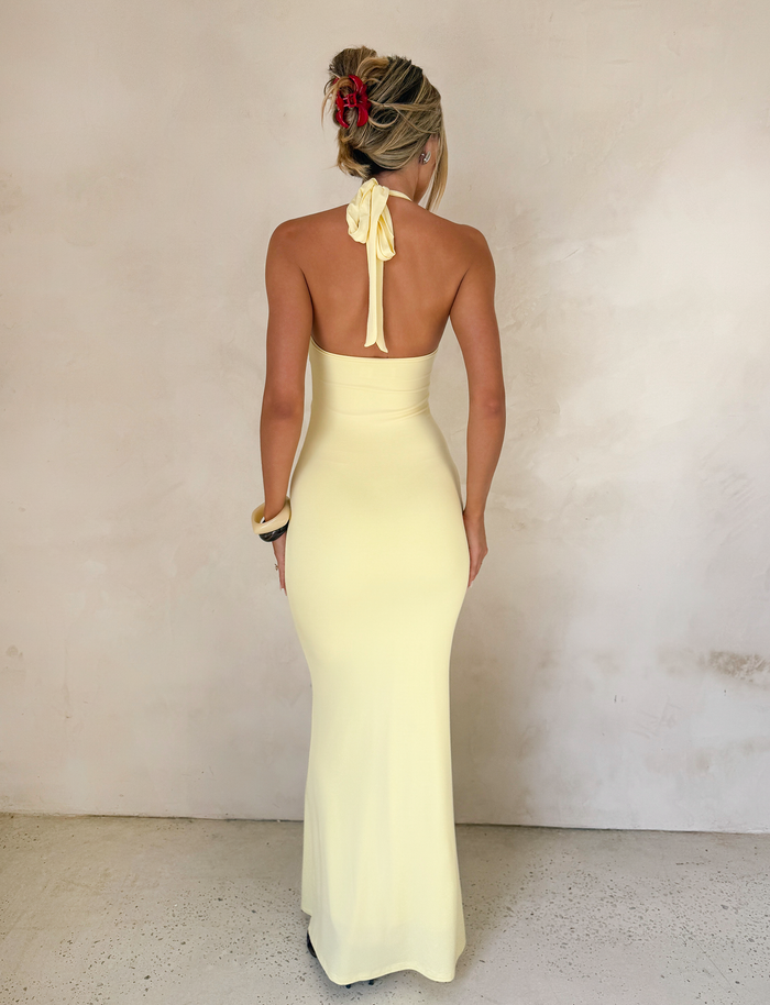 A woman with blonde hair in an updo, accessorized with hair clips and a tortoise shell ring, models the TIGERMIST ZENEVA MAXI DRESS in Lemon—a backless halter style—as she faces a light textured wall with her back to the camera.
