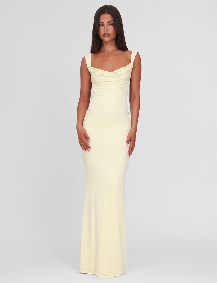 A woman with long brown hair wears the TIGERMIST ZITA MAXI DRESS - LEMON, a pastel yellow, floor-length, sleeveless gown with a draped neckline, standing against a plain light gray background.