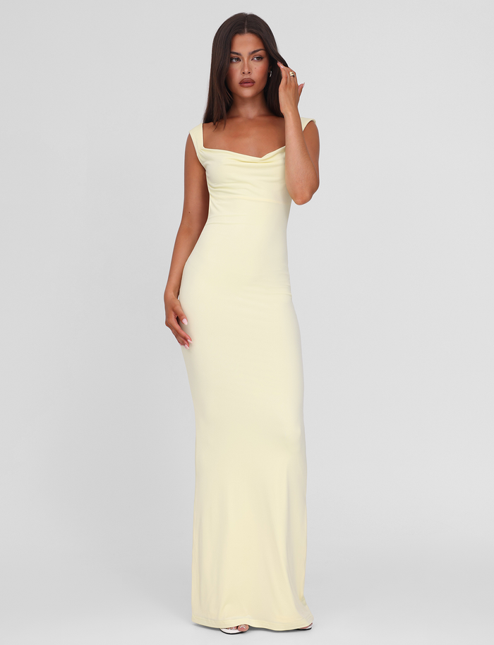 A woman with long brown hair wears the sleeveless, floor-length ZITA MAXI DRESS - LEMON by TIGERMIST, featuring a draped neckline, standing against a plain light gray background.