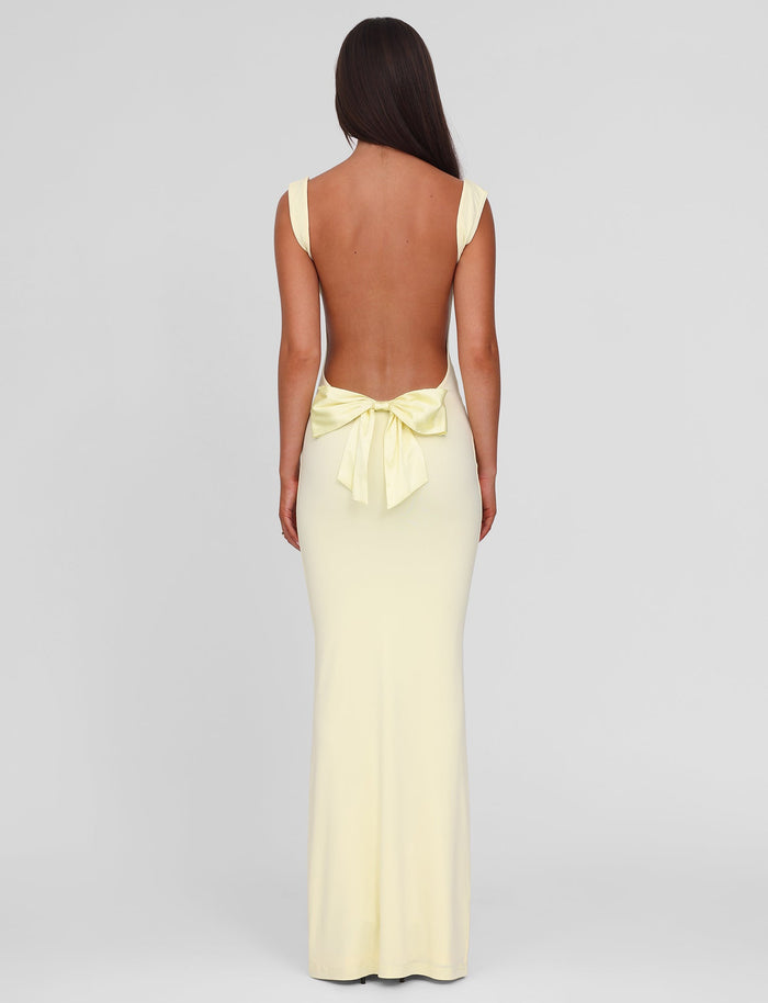 A woman with long dark hair stands facing away in the TIGERMIST ZITA MAXI DRESS - LEMON, a pastel yellow, sleeveless, backless bodycon dress featuring a large bow at the lower back. The background is plain light gray.